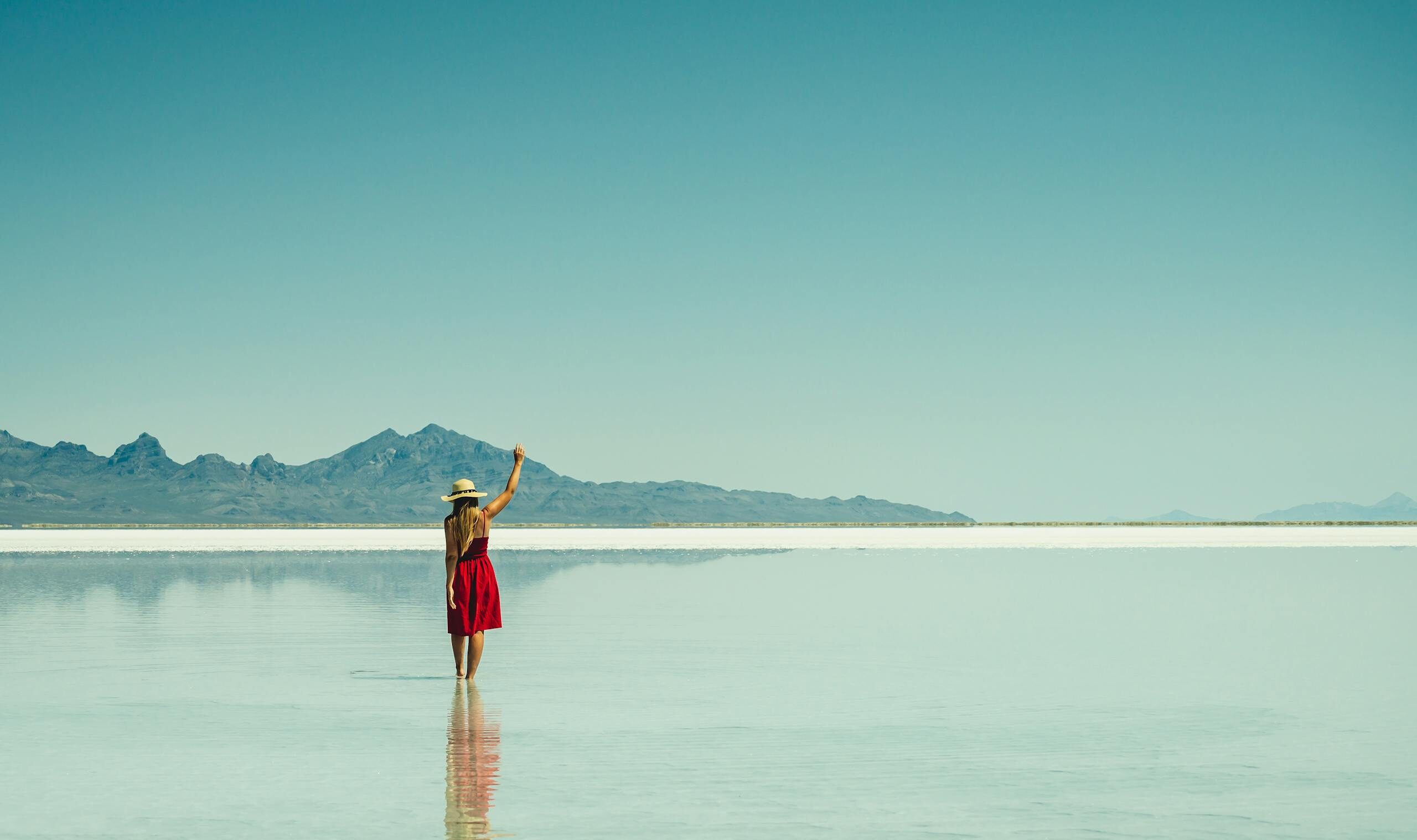 A woman in a red dress stands at a serene salt flat with mountains in the background, enjoying solitude and reflection.