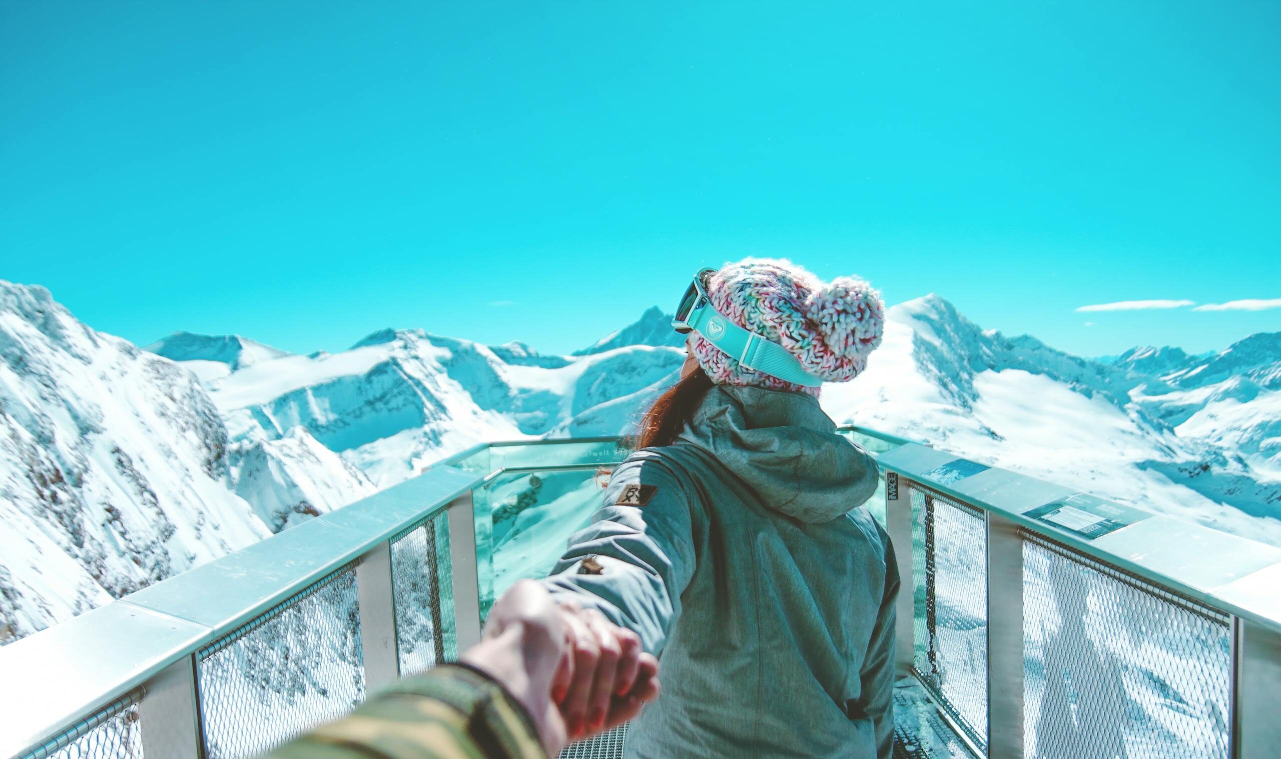 A woman in winter clothing holds a hand and walks on a scenic snow-covered mountain deck in Zell am See.